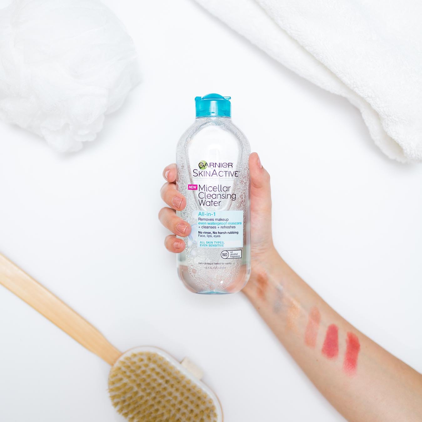 Garnier SkinActive Micellar Cleansing Water All-in-1 Waterproof 13.5 oz with blue lid being held by a female hand with 6 shades of makeup swiped across her arm over a white background beside a wooden long-handled scrub brush, folded white bath towel, and a white pouf.